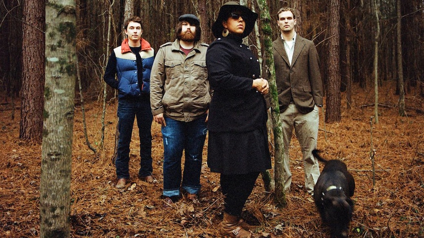 Four members of Alabama Shakes stand in the woods wearing jackets. A brown dog is by their side.