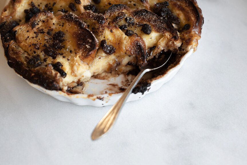 A spoon is tucked into a large bread and butter pudding after a portion has been removed, a winter dessert recipe.