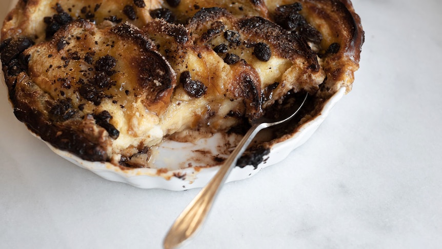 A spoon is tucked into a large bread and butter pudding after a portion has been removed, a winter dessert recipe.
