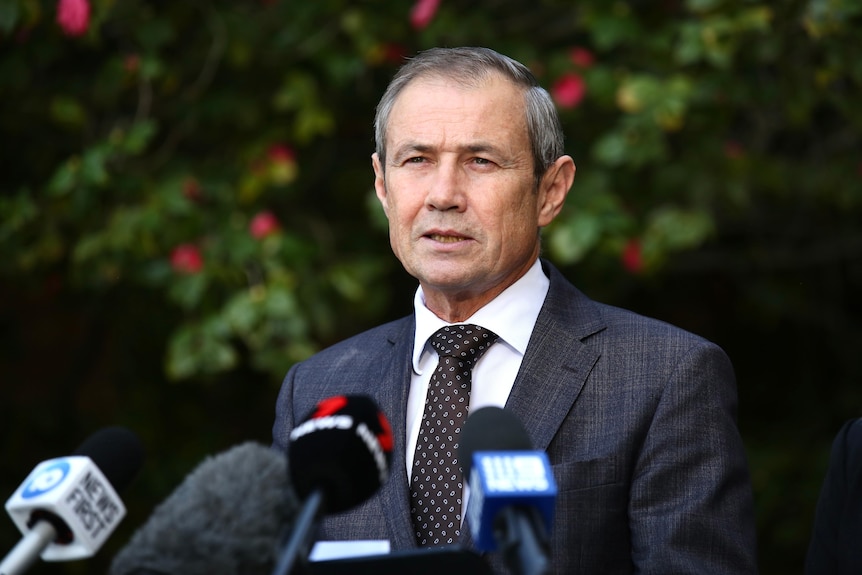 A man in a suit with a dark tie speaks in front of microphones. 