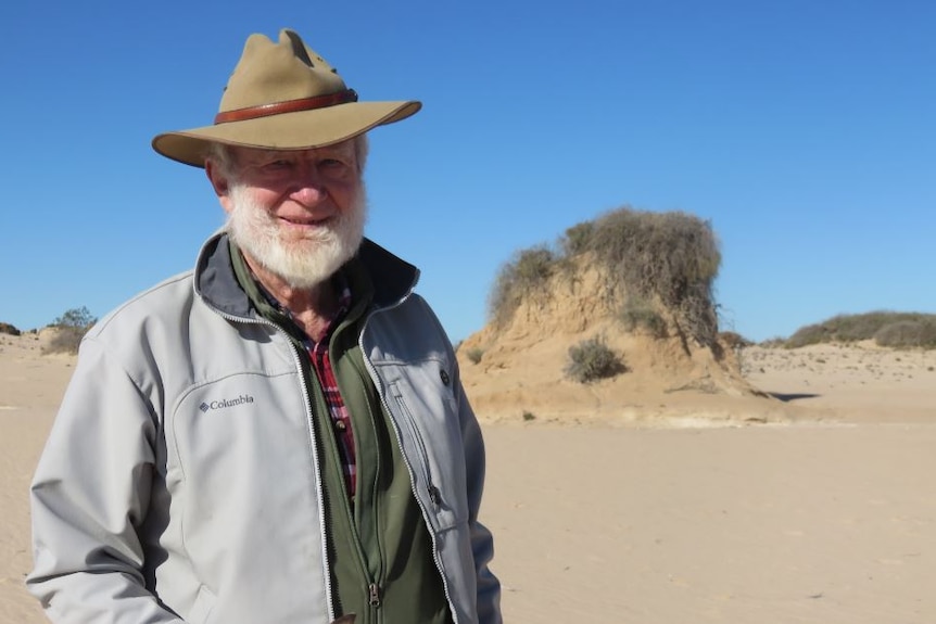An elderly man with a white beard and brimmed hat stands in front of a sand-covered landscape