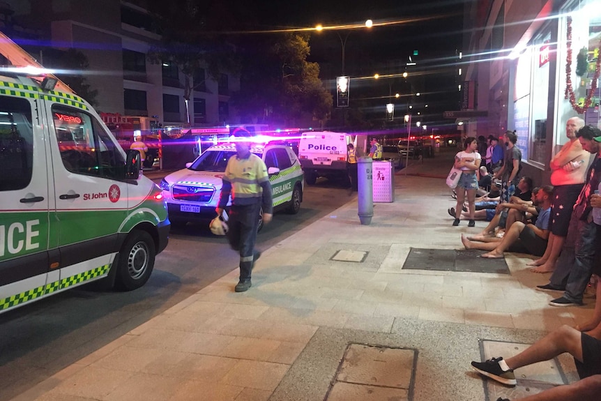 A line of people sit on a footpath next to some ambulances and fire trucks.