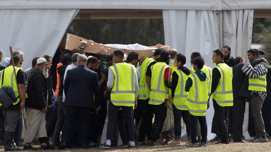 People wearing high visibility help carry a cardboard coffin containing a body wrapped in a white shroud.