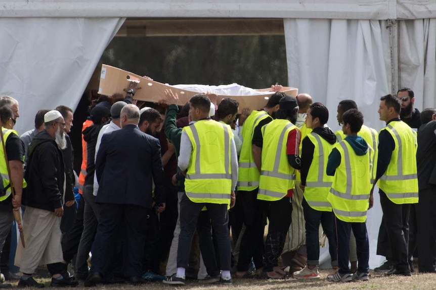 A group of men carry a cardboard coffin containing a body wrapped in a white shroud.