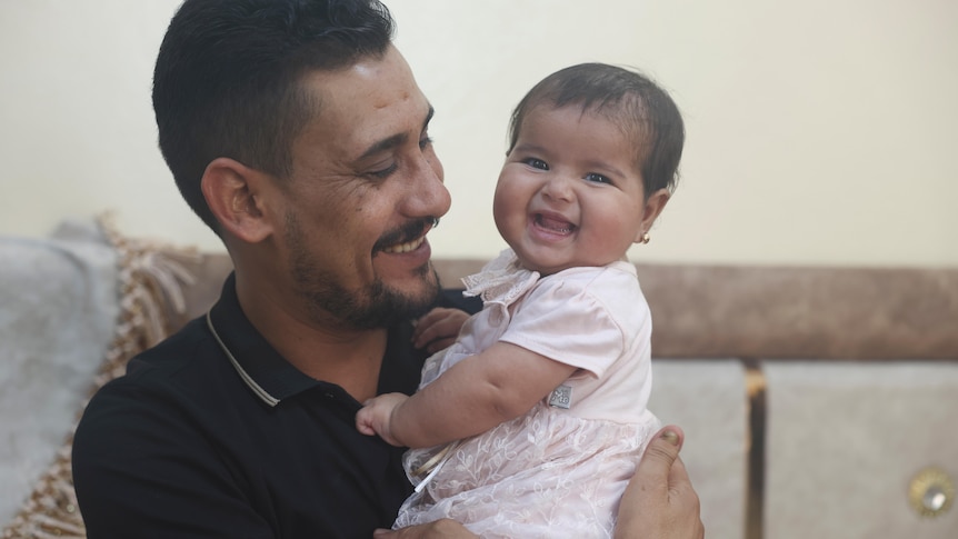 A smiling man holds a smiling 6-month-old baby