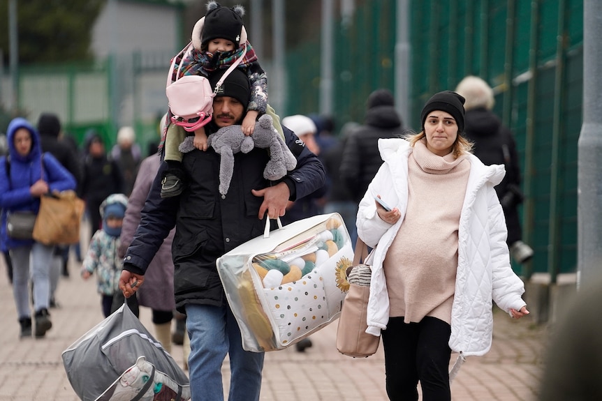 a child sits on the shoulders of a man carrying bags with a woman walking next to him as other refugees can be seen behind