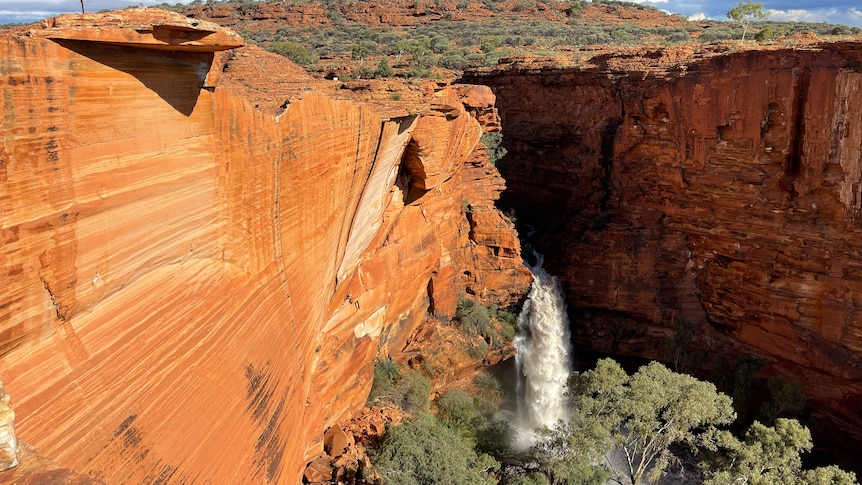 ochre rockface with gushing white water falling below into a water hole with green trees around.