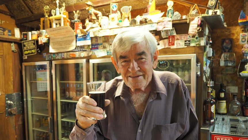 Gentleman with white hair holding glass and smiling up to camera with trophies and bar fridges in background