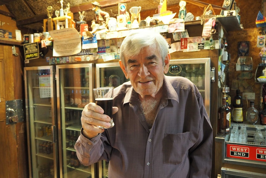 Gentleman with white hair holding glass and smiling up to camera with trophies and bar fridges in background