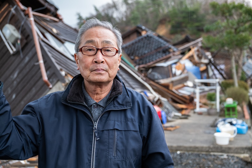 Image of an older man with glasses wearing a blue raincoat. Behind him are houses that have been destroyed.