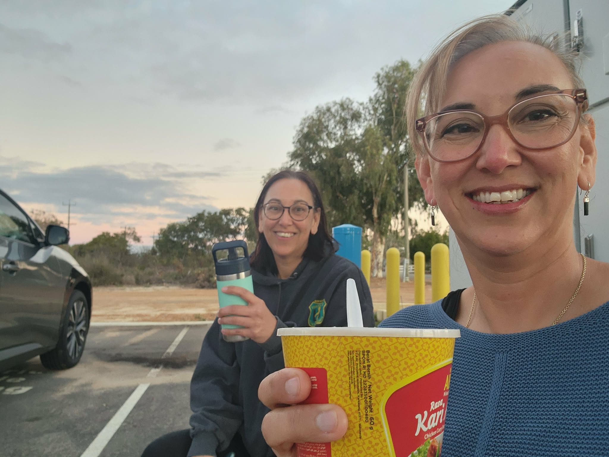 Two women smiling at the camera as they sit near a car with trees in the background.