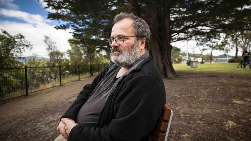 Middle aged man with beard and glasses sits on park bench near large tree.