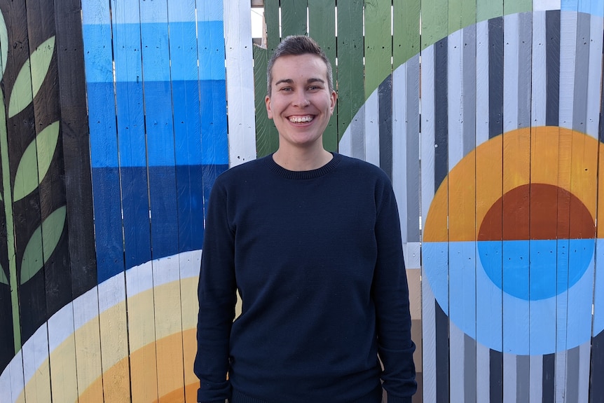 Steff Fenton wearing navy, long-sleeved shirt, standing in front of colourfully painted fence.