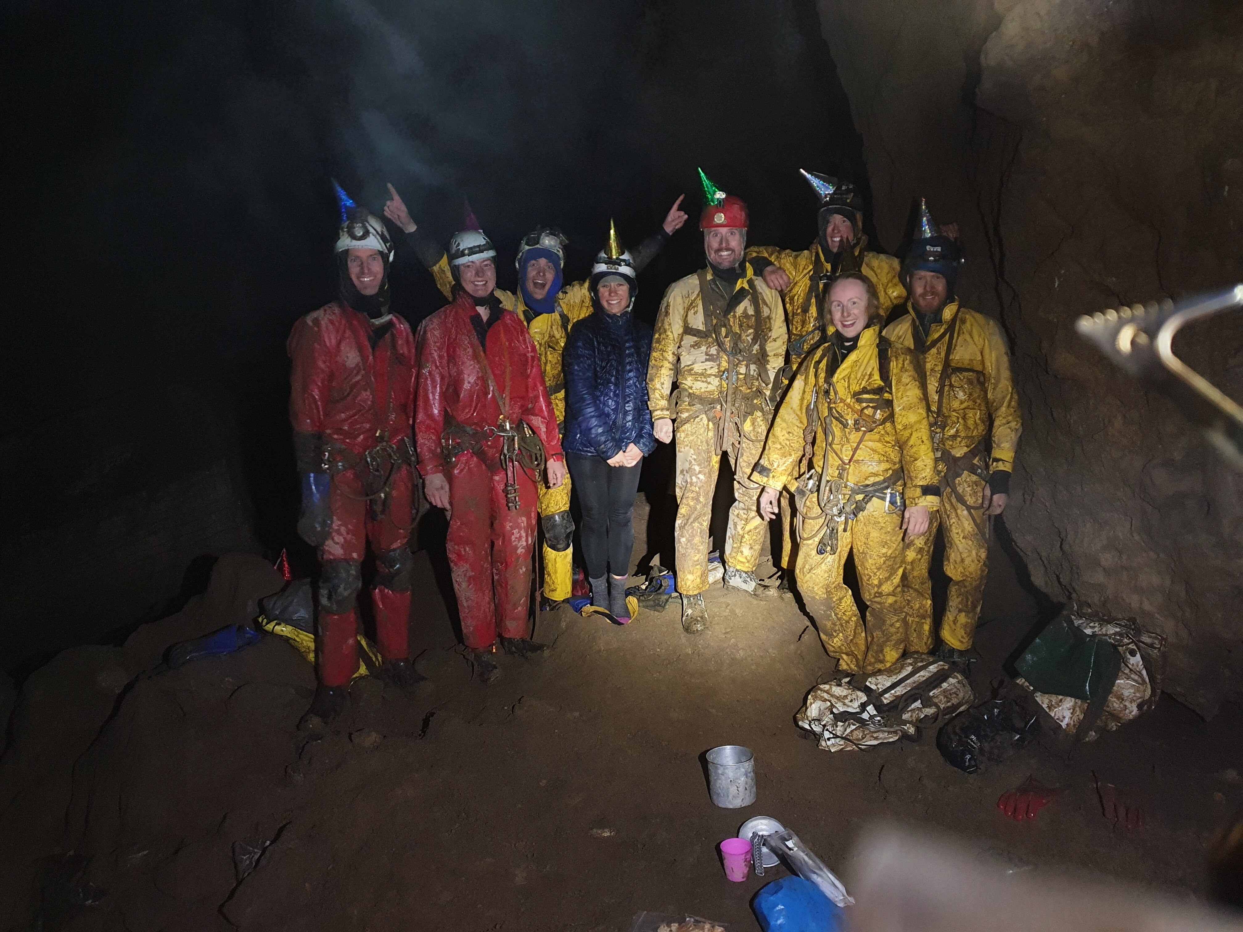 A group of people smiling underground, wearing muddy overalls and helmets. 