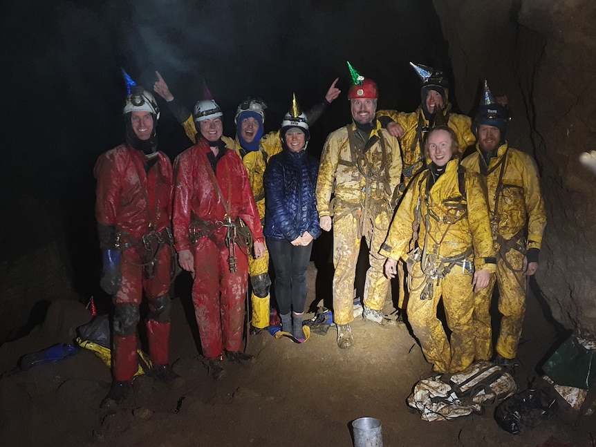 A group of people smiling underground, wearing muddy overalls and helmets. 