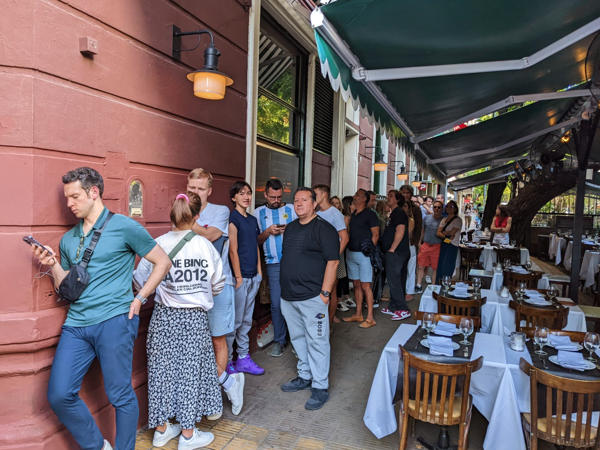 A line of people stand outside a resturant next to ourdoor seating. 
