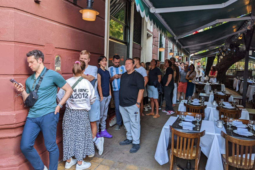 A line of people stand outside a resturant next to ourdoor seating. 