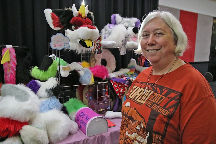 A woman with white hair smiles at the camera while standing in front of a stall with furry animal faces and ears