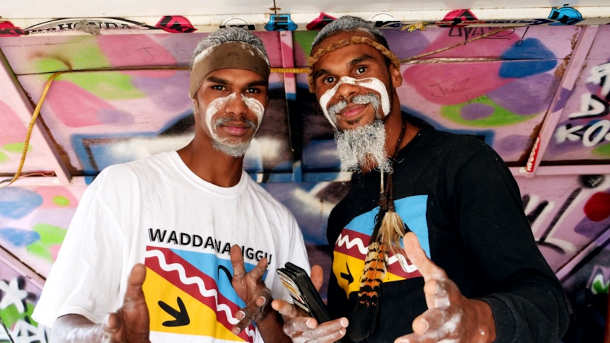 two men with traditional face painting stand in a colourful van looking at the camera