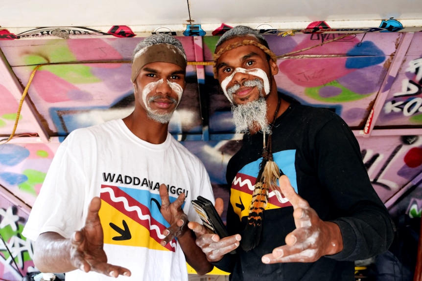 two men with traditional face painting stand in a colourful van looking at the camera