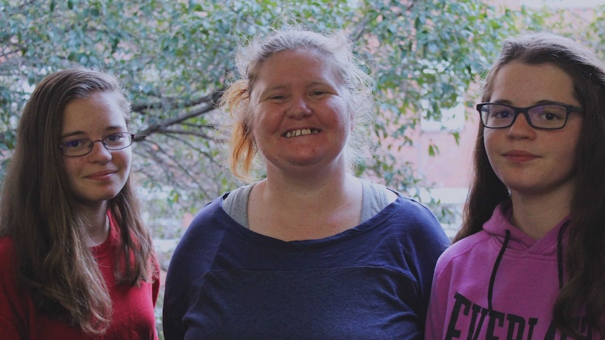 Amanda White is smiling and standing between her two twin daughters who are 14 years old. Both daughters have glasses on.