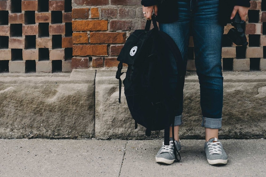 A young person stands against a wall holding a backpack to depict the pressure teens experience when finishing high school.