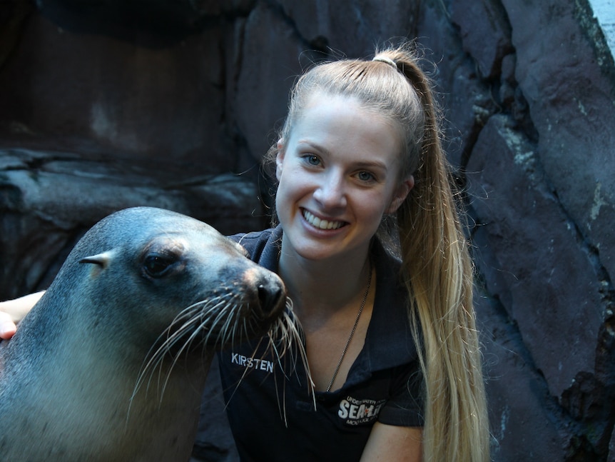Sea lion and young lady sit together. Lady is smiling.