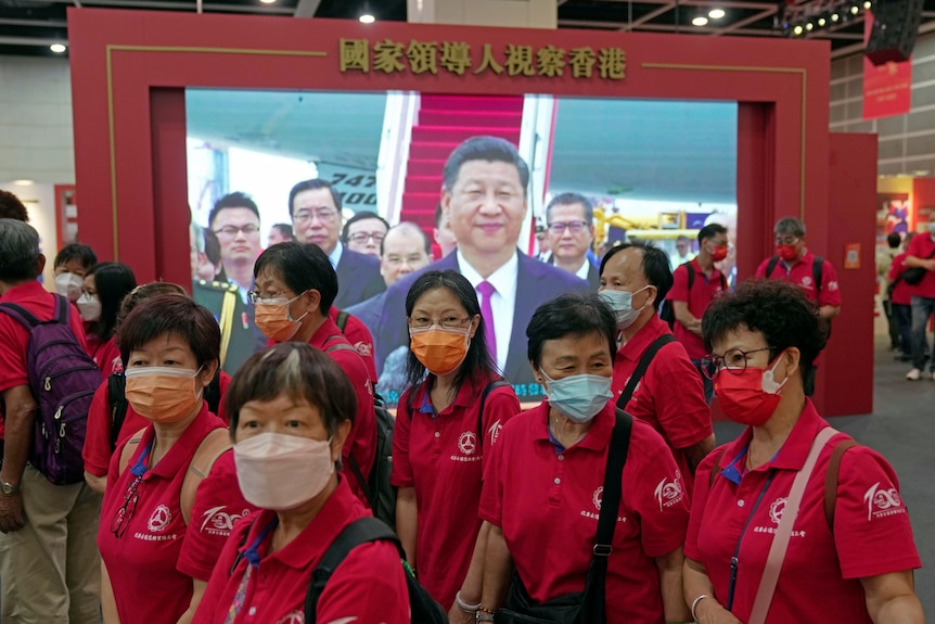 A group of women in red shirts and face masks walk past a screen showing Xi Jinping