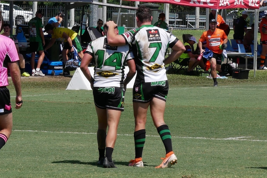 Two male football players, standing in white and green football jerseys on green grass 