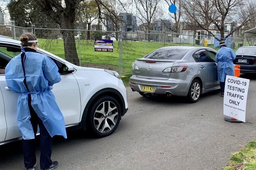 Health staff at cars in a queue near a COVID-19 testing sign.