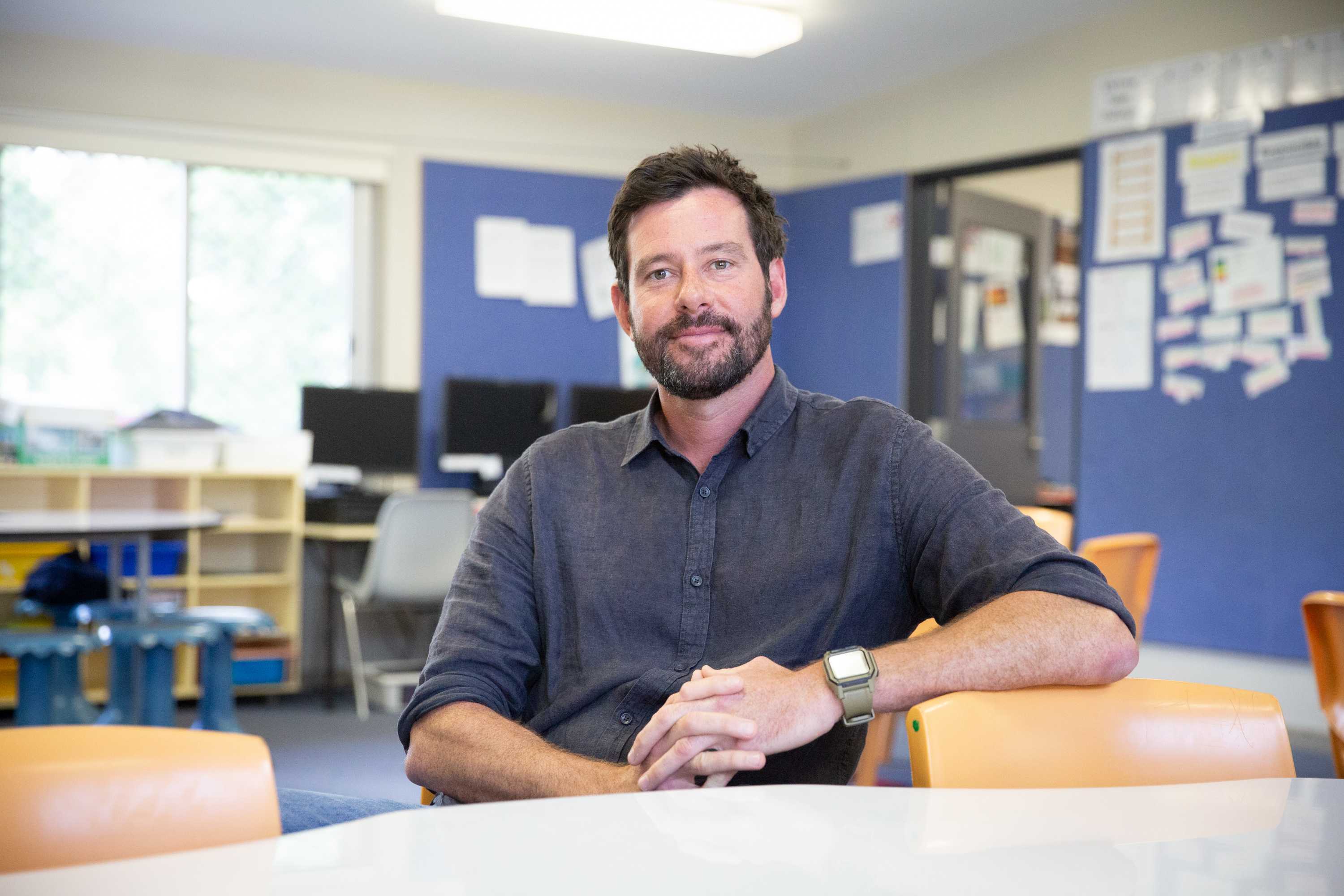 A man wearing a black collared shirt with no tie sitting in a classroom with his left arm leaning on the back of a chair