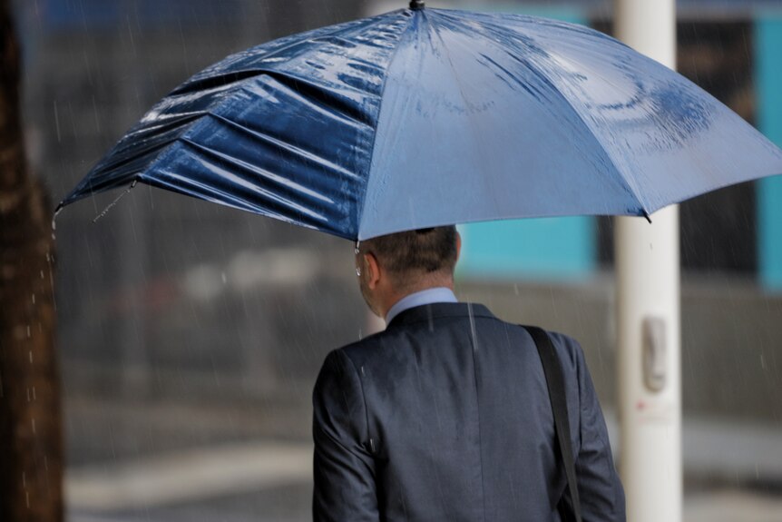 A man in suit walks with a broken umbrella in pouring rain.