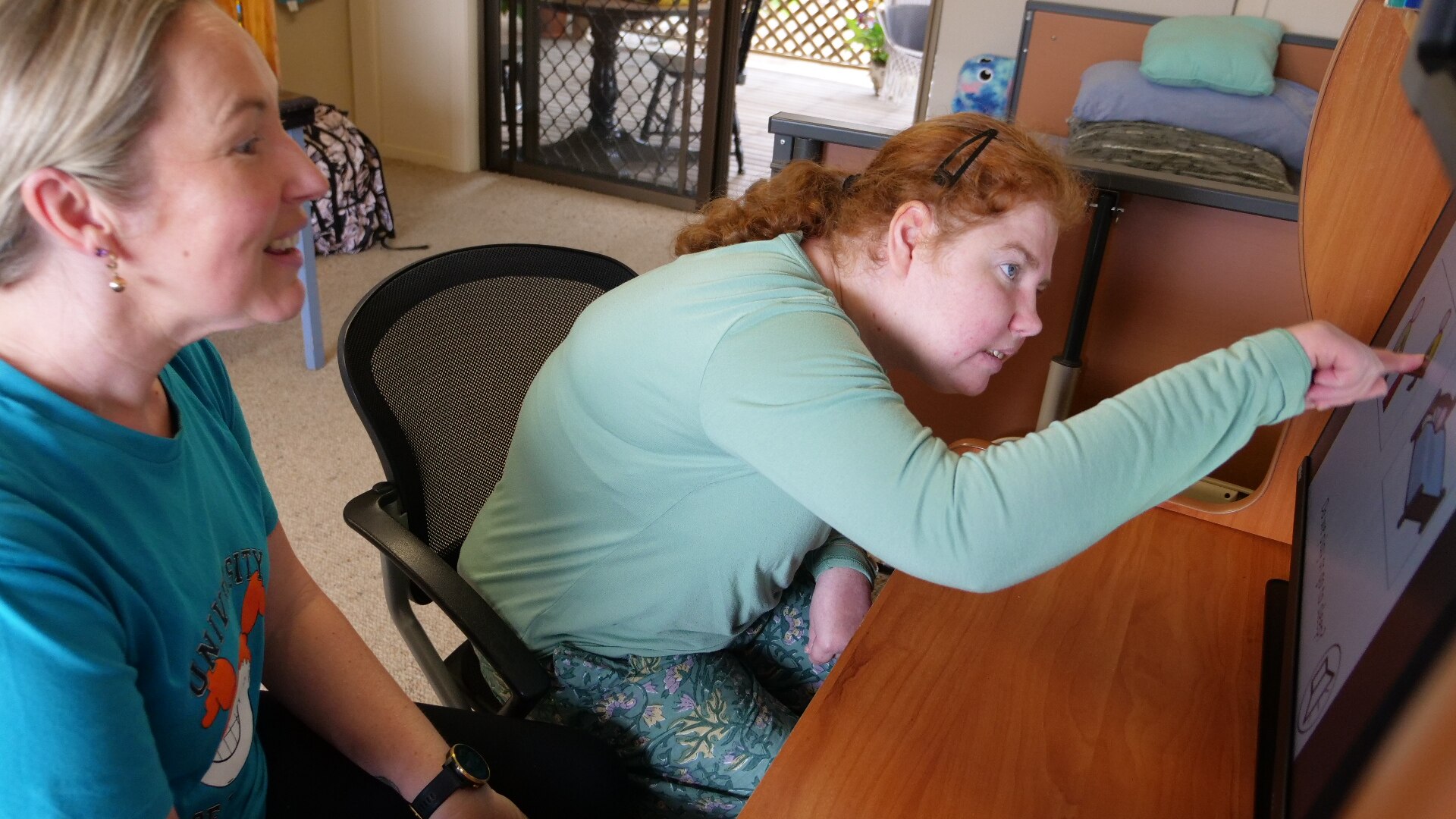 A woman with a disability sitting at her computer pointing to the screen with her support worker smiling beside her 