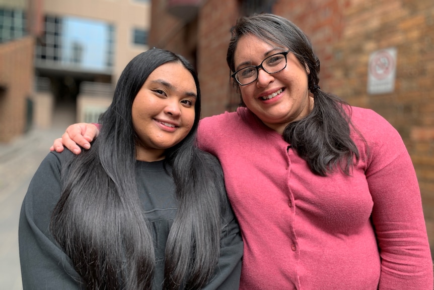 A woman in glasses and pink jumper, similes and holds a smiling young girl with long black hair and a black jumper