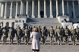A woman wearing a grey coat stands in front of a group of men in uniforms and wearing helmets and masks in front of the Capitol.