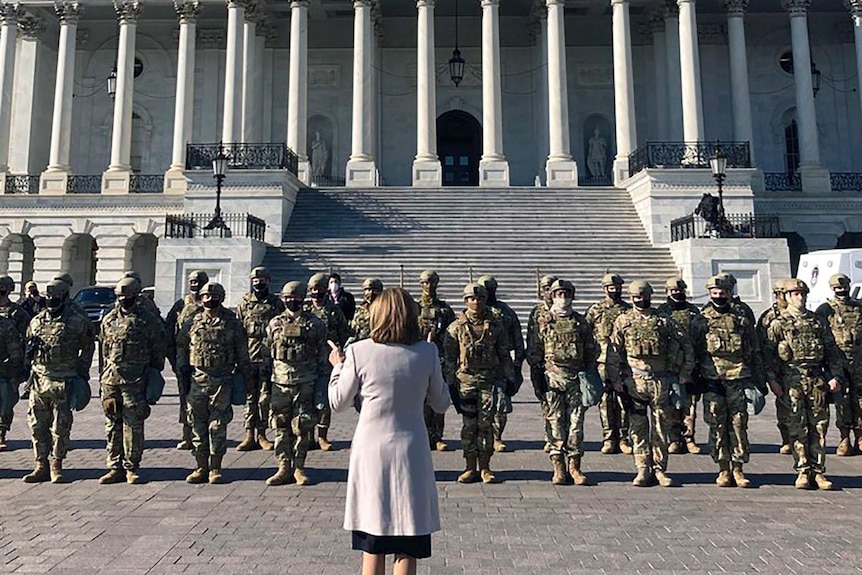 A woman wearing a grey coat stands in front of a group of men in uniforms and wearing helmets and masks in front of the Capitol.