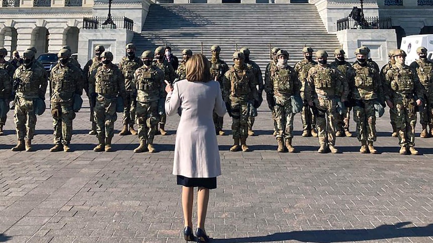 A woman wearing a grey coat stands in front of a group of men in uniforms and wearing helmets and masks in front of the Capitol.