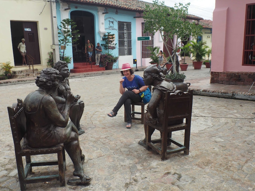 A woman in a pink hat sitting among three statues of men sitting down in a colourful stree square in Asia