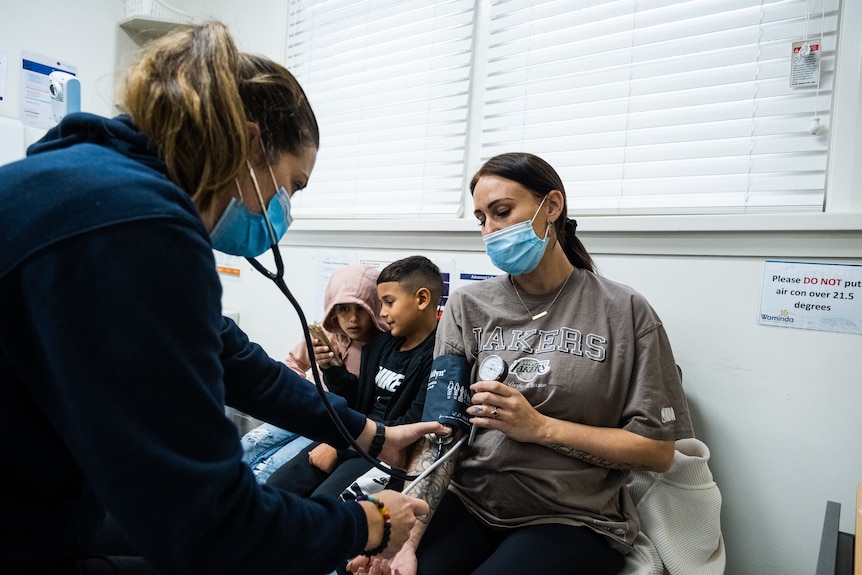 A midwife wearing a facemask checks the blood pressure of a female patient while her children sit next to her. 