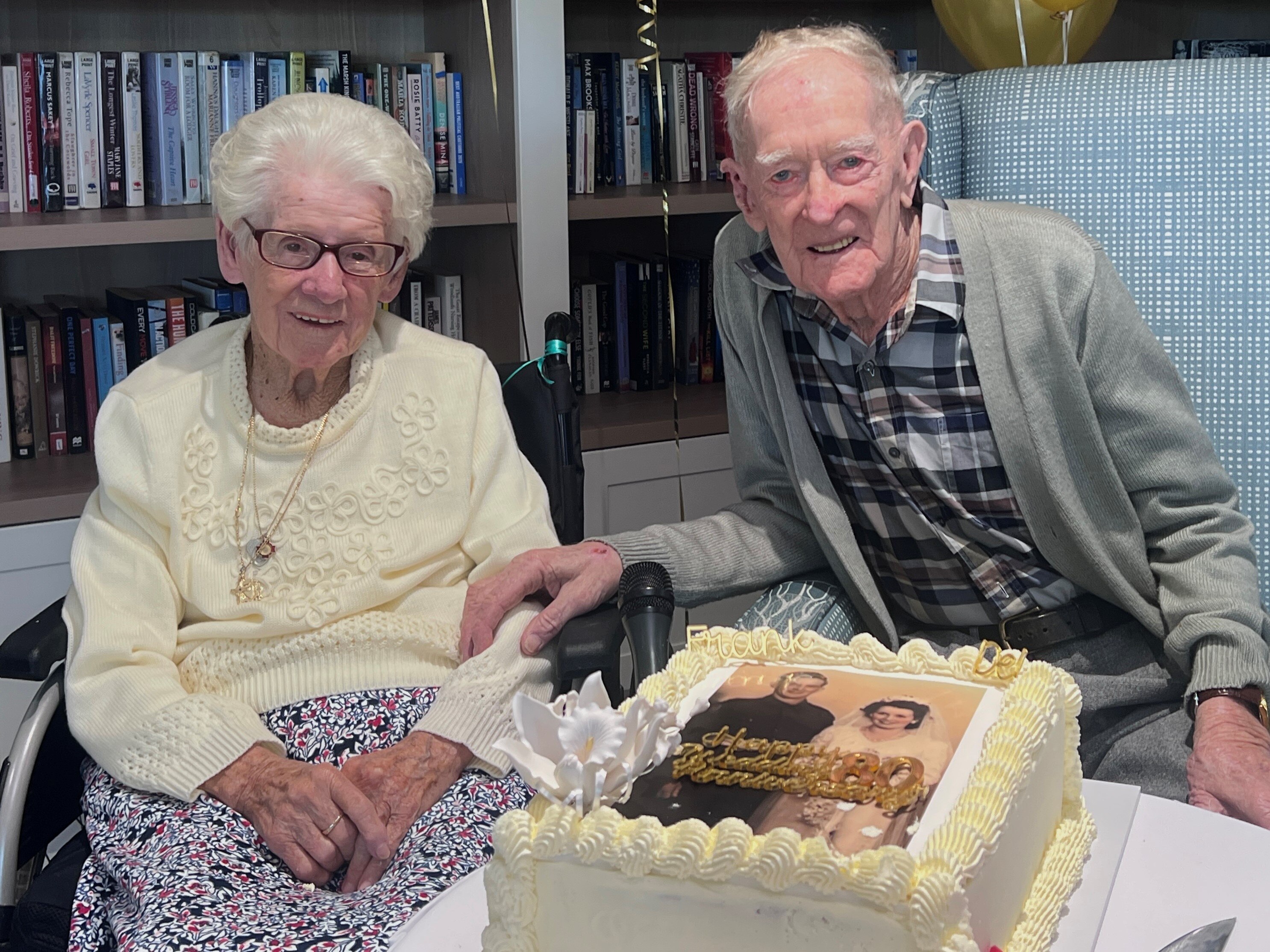Frank and Delma sit side-by-side, holding hands, in front of an anniversary cake, decorated with their wedding photo. 