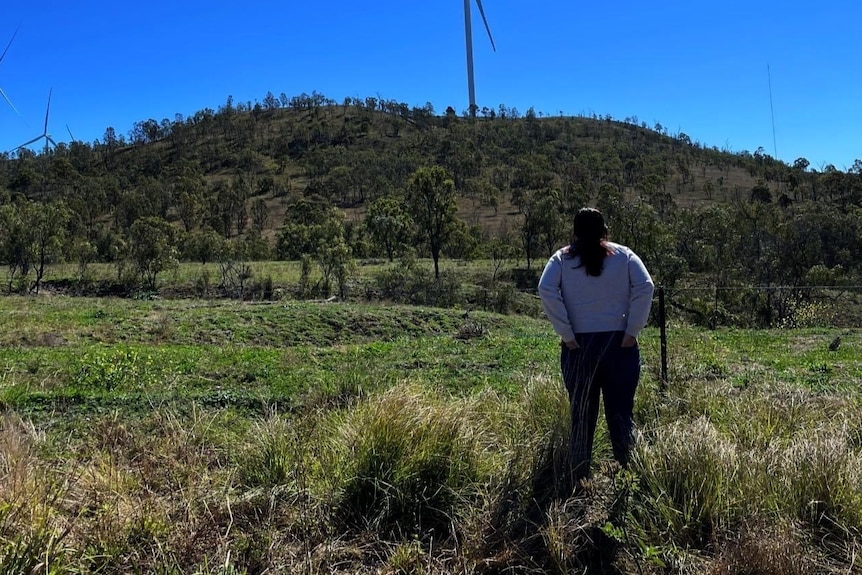 A woman stands in a field with her hands in her back pockets staring at the mountain in front of her.