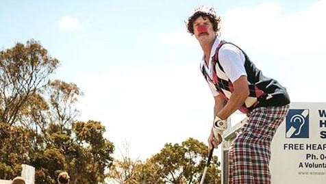 A clown on stilts gets ready to hit a pink golf ball with a very long golf club.