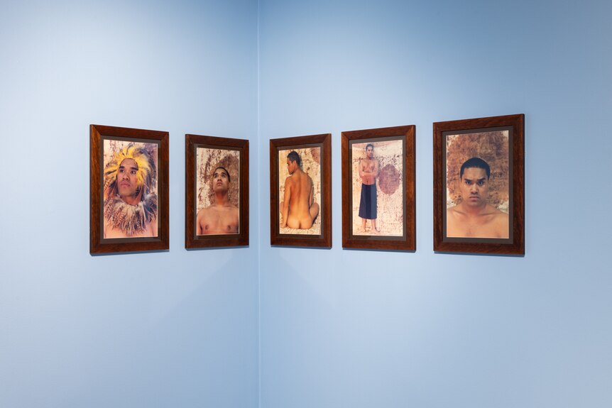 Five arts works in brown frames hang on blue walls.