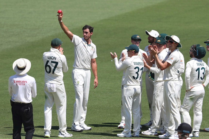 Mitchell Starc holds the pink ball in his hand above his head as his teammates stand around him and clap