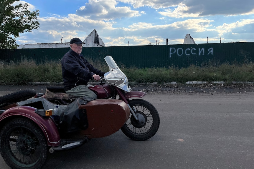 Un uomo guida la sua moto davanti a un cartello che dice in russo "Russia" Nella città restaurata di Lyman.