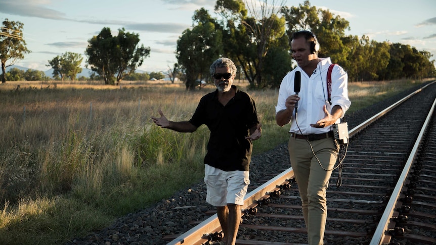 Allan wearing headphones and talking into microphone walking along train tracks with Craigie.