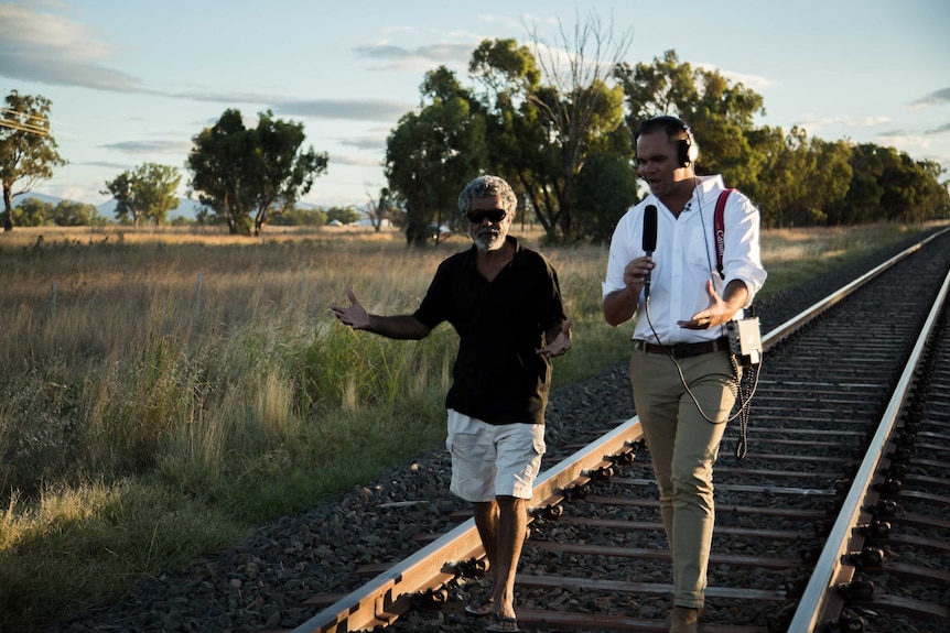 Allan wearing headphones and talking into microphone walking along train tracks with Craigie.