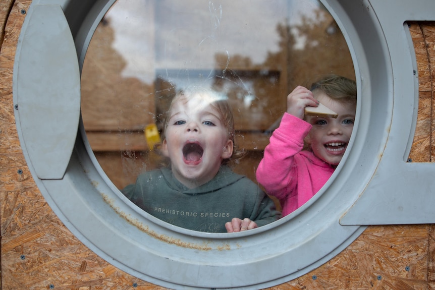 Twins Billie and August play behind a glass window.