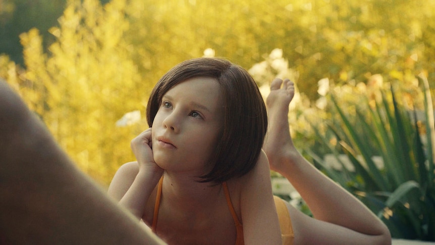 A young AI girl sitting outside in bathers in the film The Trouble with Being Born
