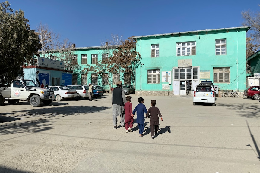 Un homme et trois enfants marchent vers un bâtiment de l'hôpital bleu vif